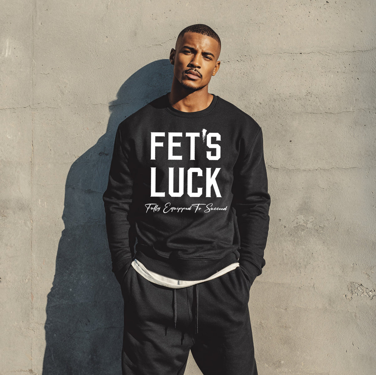 Male model wearing a black GET-ILL FET'S LUCK graphic crewneck sweatshirt with bold white lettering against an urban concrete wall.