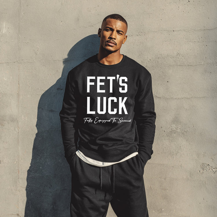 Male model wearing a black GET-ILL FET'S LUCK graphic crewneck sweatshirt with bold white lettering against an urban concrete wall.