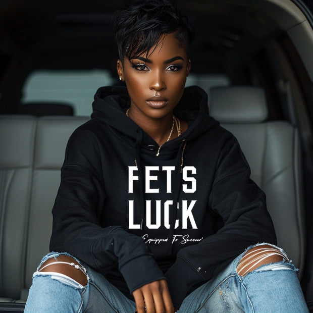 Female model seated in an urban setting wearing a black GET-ILL FET'S LUCK graphic hoodie with bold white lettering.
