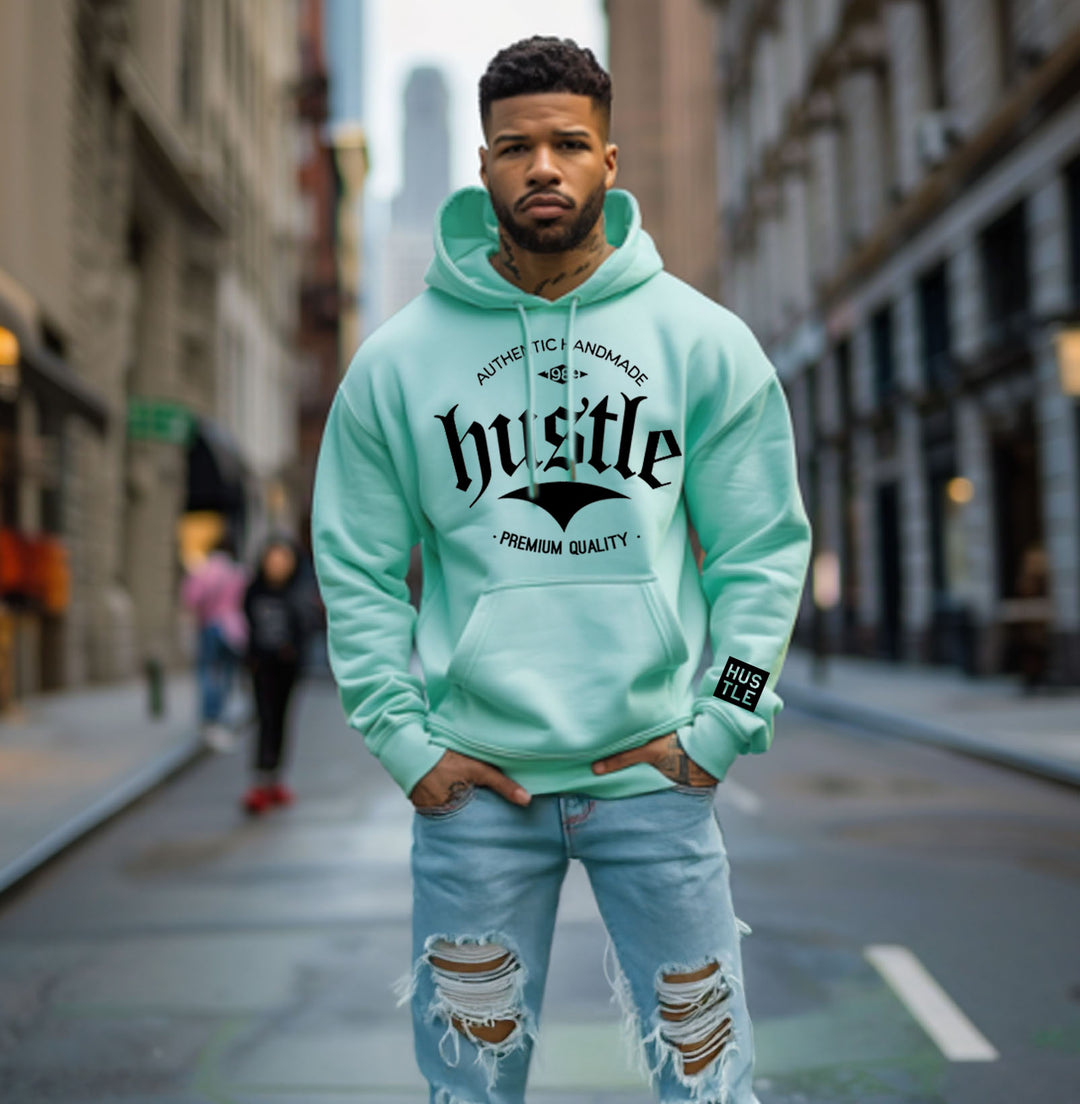 Close-up of light green 
hoodie on a person, showing the "hustle" graphic with "AUTHENTIC HANDMADE" and "PREMIUM QUALITY" text.
