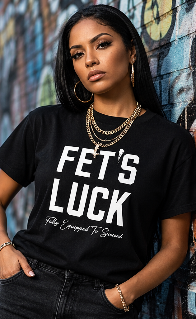 Female model wearing GET-ILL FET'S LUCK black graphic T-shirt with bold white lettering, styled with gold jewelry against a graffiti wall.